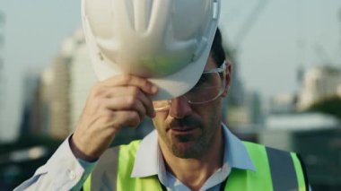 Koruyucu gözlüklü insan mühendisinin portresi kameraya bakarken kaskını takıyor. Yansıtıcı yelek giyen erkek müteahhit inşaat görevlerine hazırlanmak için sert başlık ayarlar