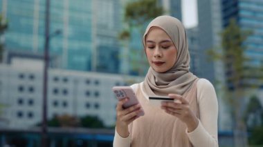 Başörtüsü takan Müslüman kadın portresi internette kredi kartı ve akıllı telefon kullanıyor. Malezyalı kadın şehir ortamında bankacılık uygulaması yoluyla parayı güvenli bir şekilde transfer ediyor