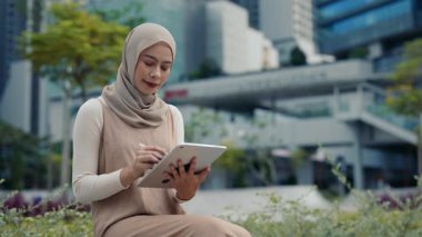 Genç Müslüman iş kadınının portresi şehir parkında bulunan tablet cihazına dijital kalem kullanıyor. Türbandaki Malezyalı bayan grafik tasarımcısı şehir ortamındaki uzak çalışmaları kucakladı