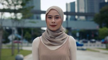 Başörtülü Müslüman iş kadını portresi şehir parkındaki kamera standlarına bakıyor. Geleneksel kıyafetler giyen Malezyalı güzel bir bayan ofis binalarına karşı sokakta poz veriyor.