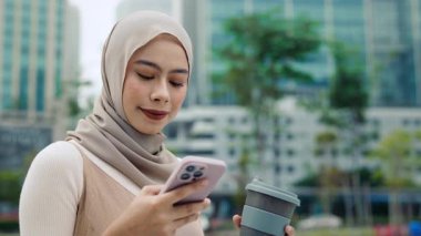 Genç Malezyalı tesettürlü iş kadını portresi açık havada kağıt kahve fincanı tutuyor. Güzel Müslüman kadın modern şehirde cep telefonundan mesaj okuyor. Şehir hayatı