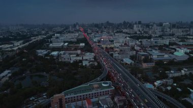 Bangkok 'ta yüksek bir otoyoldaki yoğun trafiğin gece görüşü. Durağan bir görüntüde insansız hava aracı tarafından görüntülendi. Arka planda şehrin ufuk çizgisine doğru uzanan kırmızı arka farlar görülüyor..