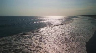Endonezya, Bali 'deki kumlu sahil şeridine düşen okyanus dalgalarının drone görüntüsü, su yüzeyine yansıyan güneş ışığı.