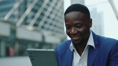 Afro-Amerikalı iş adamının portresi, kentsel arkaplanda dijital tablet ekranına bakarken gülümsüyor. Genç yönetici, şehir sokaklarındaki cihazla iş verilerinin büyümesini çevrimiçi olarak analiz ediyor