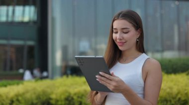 Beyaz kolsuz tişörtlü kadın portresi dijital tablet üzerinde çalışıyor ve şehir sokaklarında gülümsüyor. Neşeli kadın parşömenleri portatif bilgisayarla İnternet üzerinden halka açık bahçede duruyor.