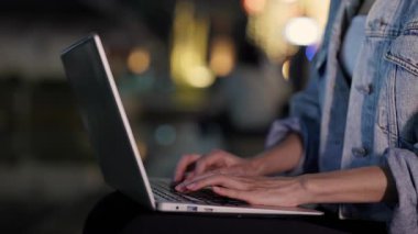 Kadın eli görüntüsü şehir merkezinde dizüstü bilgisayarda klavye kullanıyor. Kadın uzaktan kumandalı çalışan akşam kasabasında oturan taşınabilir bilgisayara rapor yazıyor. Dijital teknoloji