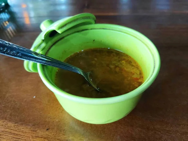 Indonesian special liquid chili sauce.  served in a dirty green plastic container.  equipped with a spoon on a wooden table.  Usually eaten with meatballs or soup