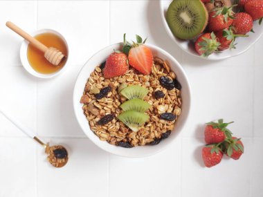 A bowl of granola. Completed with dried fruit, kiwi, strawberry, honey, honey dipper, mug jug, spoon. Healthy food concept. Isolated background in white.