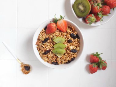 A bowl of granola. Completed with dried fruit, kiwi, strawberry, honey, honey dipper, mug jug, spoon. Healthy food concept. Isolated background in white.