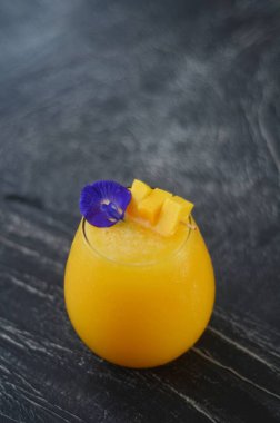 Bir bardak mango suyu. Üstünde mango dilimleri ve bezelye çiçekleri var. Sağlıklı ve tropikal içecekler. karanlık ruh hali fotoğrafçılık konsepti.