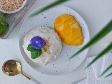 Mango pilavı. Tayland 'dan gelen otantik bir sokak yemeği. Yapışkan pirinç, hindistan cevizi sütü, tuz, şeker, mango ve susam tohumundan yapılmış. Beyaz tabakta servis edilmiş. Nane yapraklarıyla kaplı, bezelye çiçeği