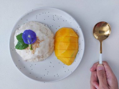 Mango pilavı. Tayland 'dan gelen otantik bir sokak yemeği. Yapışkan pirinç, hindistan cevizi sütü, tuz, şeker, mango ve susam tohumundan yapılmış. Beyaz tabakta servis edilmiş. Nane yapraklarıyla kaplı, bezelye çiçeği