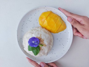 Mango pilavı. Tayland 'dan gelen otantik bir sokak yemeği. Yapışkan pirinç, hindistan cevizi sütü, tuz, şeker, mango ve susam tohumundan yapılmış. Beyaz tabakta servis edilmiş. Nane yapraklarıyla kaplı, bezelye çiçeği