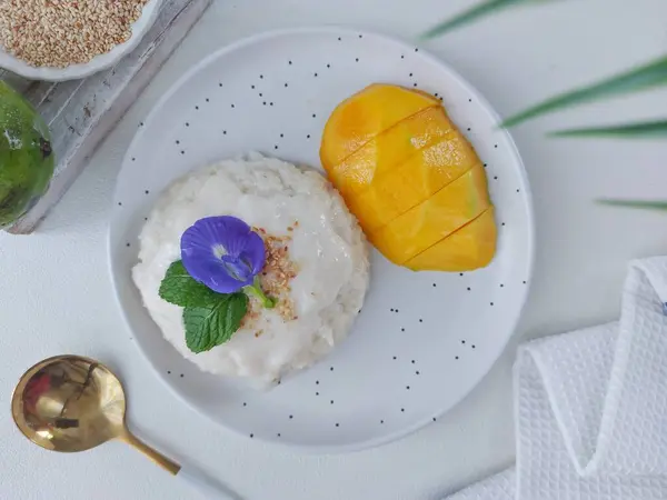 Mango sticky rice. It is authentic street food from thailand. Made from sticky rice, coconut milk, salt, sugar, mango, sesame seed. Served on white plate. Topped with mint leaves, butterfly pea flower