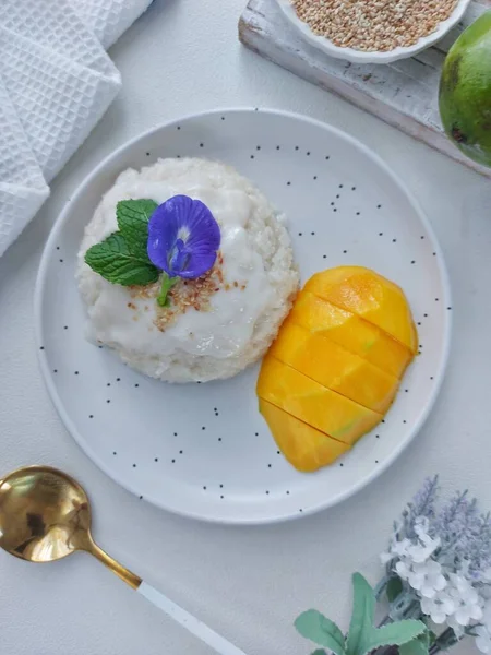 Mango sticky rice. It is authentic street food from thailand. Made from sticky rice, coconut milk, salt, sugar, mango, sesame seed. Served on white plate. Topped with mint leaves, butterfly pea flower