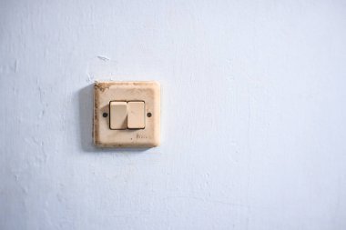 Eski, yıpranmış bir ışık düğmesi, dokulu bir duvara monte edilmiş. Düğme krem rengi plastikten yapılmış ve iki düğmesi var. Düğmedeki leke ve kir ona klasik ve kırsal bir görünüm kazandırıyor. Bu görüntü nostalji hissi uyandırıyor.