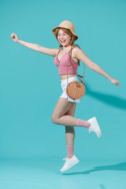  Çok heyecanlı, mutlu Asyalı bir kadın, parlak turkuaz arka planda, yaz kıyafetiyle, şapkasıyla zıplıyor.