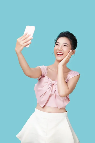 快乐的年轻女人一边做傻脸 一边用装有滤镜的智能手机应用自拍 站在蓝色的背景下 — 图库照片