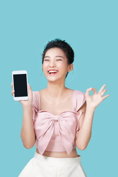 陽気アジアの女性は スマートフォンを保持し ライトシアンの背景にOkサインを示しています — ストック写真