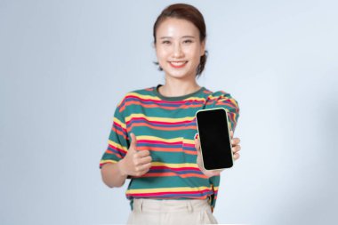  Mutlu Asyalı kız elinde akıllı telefon tutuyor ve tamam işareti gösteriyor, gri arka planda işaret veriyor.