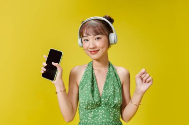 Sarı arka planda yalnız poz veren kadın kulaklıkla müzik dinliyor.