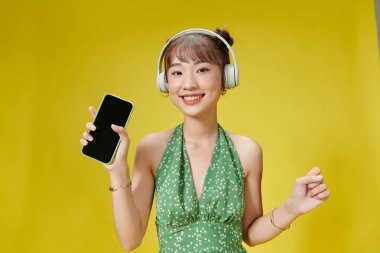 Sarı arka planda tek başına duran bir kadın, kulaklıkla müzik dinliyor, şarkı söylüyor.