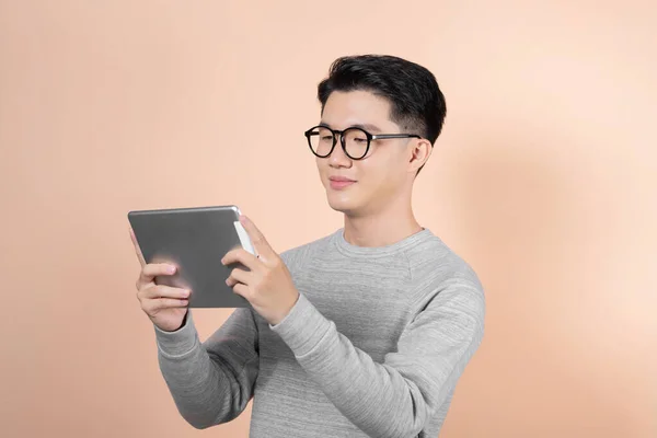 stock image Handsome young asian man holding digital tablet and looking at it with smile while standing against beige background