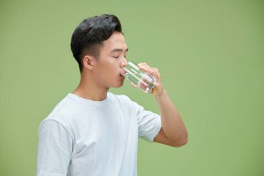 Mutlu Asyalı genç adam yeşil arka planda camdan su içiyor.