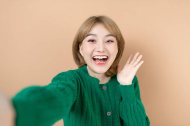 Bej arka planda izole bir şekilde poz veren neşeli, genç ve güzel bir kadının görüntüsü. Kamerayla selfie çek.