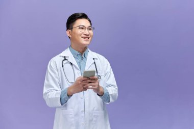 Beyaz önlüklü genç erkek doktor telefonuna bakarken gülümsüyor.