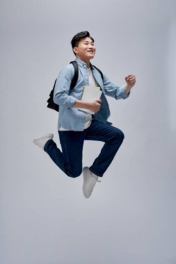 Sırt çantası taşıyan, kitap tutan, zıplayan gülümseyen Asyalı bir adamın tam boy portresi.