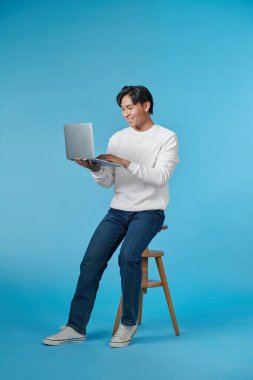 Yakışıklı duygusal adam taburede oturuyor ve mavi arka planda dizüstü bilgisayar kullanıyor.