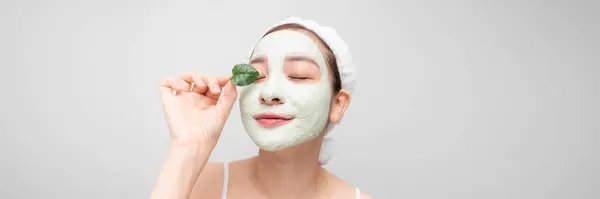 Young asian girl enjoys clay facial mask posing with a leaf on white banner.