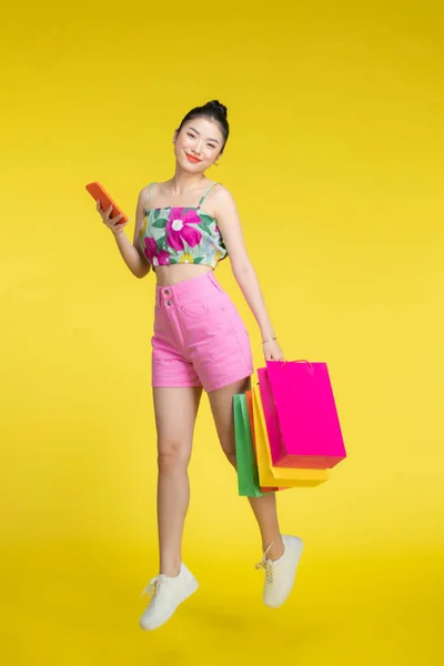 年轻精力充沛的亚洲女人拿着装有黄色背景购物袋的手机 — 图库照片