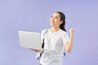 Sırt çantalı, elinde dizüstü bilgisayarla menekşe arka planda izole edilmiş mutlu kız öğrenci.