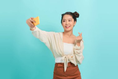 Neşeli bir kadın cep telefonu kullanıyor, selfie çekiyor ve iki parmağıyla kalp işaretini gösteriyor.