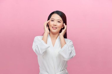 Güzel gülümseyen Asyalı kadın model, doğal makyajıyla pembe arka planda parlayan nemli cilde dokunuyor..