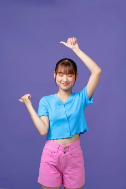 Genç Asyalı kız ayakta duruyor, eli arkada, baş parmağı havada, kendinden emin gülümsüyor.