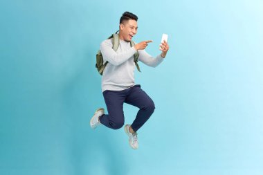 Heyecanlı Asyalı genç bir adamın portresi. Akıllı bir telefon tutuyor ve izole edilmiş açık mavi üzerine atlıyor. 