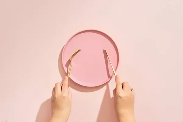 Hands hold a fork and knife over plate on pink background