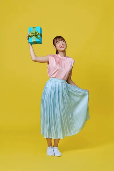 Cheerful millennial lady in pretty dress showing wrapped box with gift on yellow background