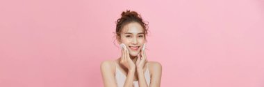 Cheerful young woman holding cotton pads while standing isolated over pink