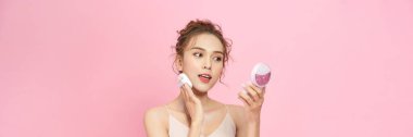 Portrait of attractive cheery girl applying blush tone foundation isolated over pink pastel background