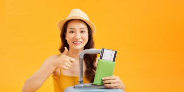 Hayalci Asyalı turist, gülümsüyor ve düşünüyor, seyahat için bavullu bilet ve pasaport tutuyor.