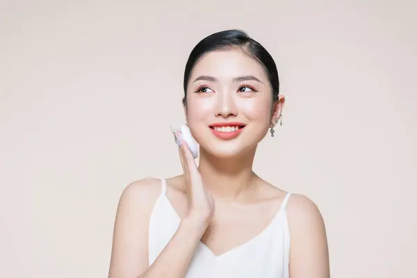 年轻的亚洲女人笑着高兴地在米色的背景上涂上浮肿的化妆品 免版税图库图片
