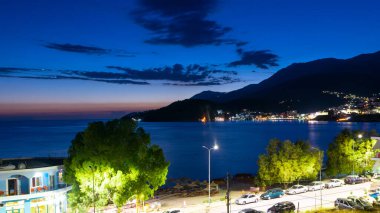 Twilight landscape of a resort Himare town located in a bay at the foot of the mountains. The village is beautifully lit by lanterns after sunset. Ionian sea. Albania.