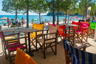 Himare, Albania - 15 July 2021: Tables and multi-colored chairs of a summer cafe on the promenade. Vacationers on beautiful clean city public beach with pine trees. Ionian Sea clipart