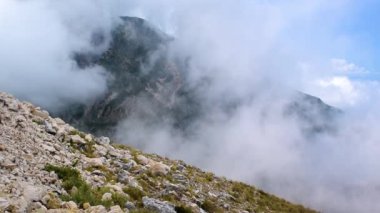 White fog slowly creeps up the high mountain. View from the highlands. A blue sunny sky is visible through the fog. Travel in Albania. Road to Llogara Pass.