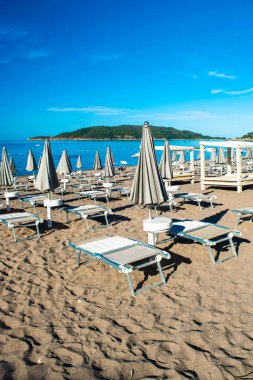 Güneşli yaz sabahında halk plajında güneş pansiyonları ve şemsiyeler. Skojl Adası geçmişi. Sakin deniz ve mavi gökyüzü. Budva Riviera. Deniz kıyısındaki tatil köyü sezonu. Becici, Karadağ.