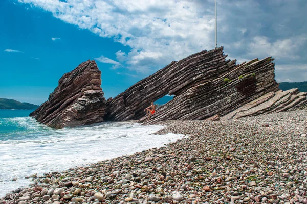 stock image Young man in front of beautiful rocks on pebble public stone beach. Rafailovici esort towns. Mountenegro. Adriatic Sea. Summer sunny day. Seaside vacation concept.